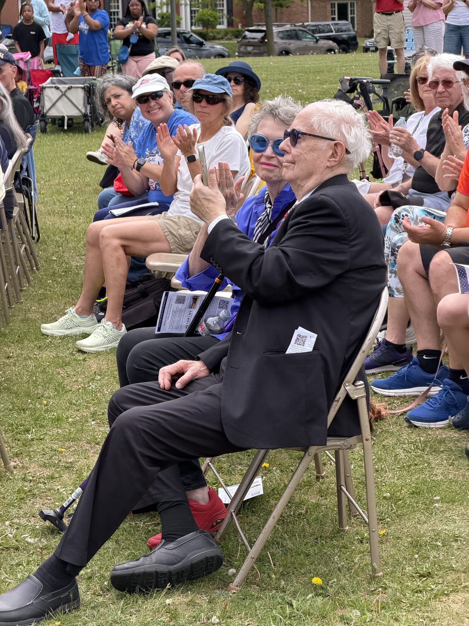 An audience member applauds during the Whitefish Bay Memorial Day service, seated on a folding chair on Armory Park's green lawn with rows of neighbors around him