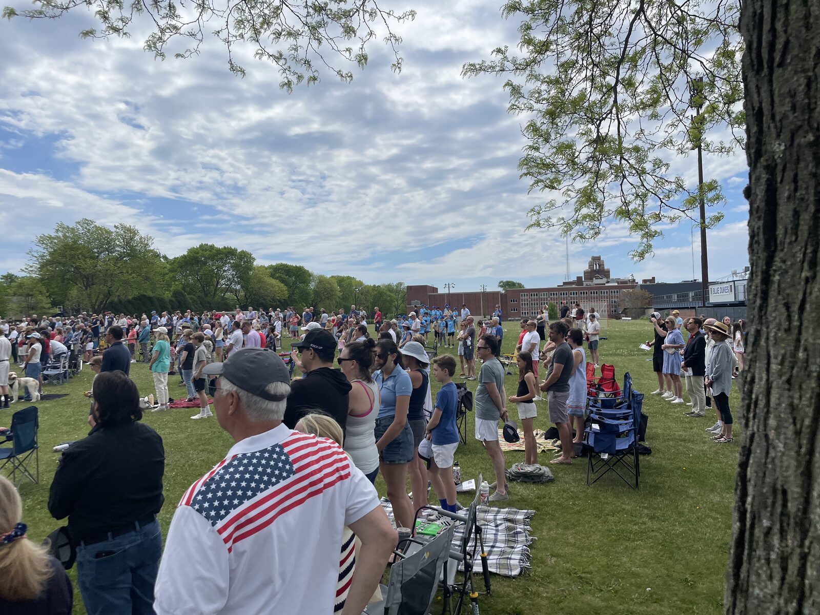 Hundreds of Whitefish Bay residents gather on the open green at Armory Park for the Memorial Day service. The school is visible in the background, families stand with picnic blankets and folding chairs, and a man in the foreground wears a polo shirt with an American flag across the shoulder.