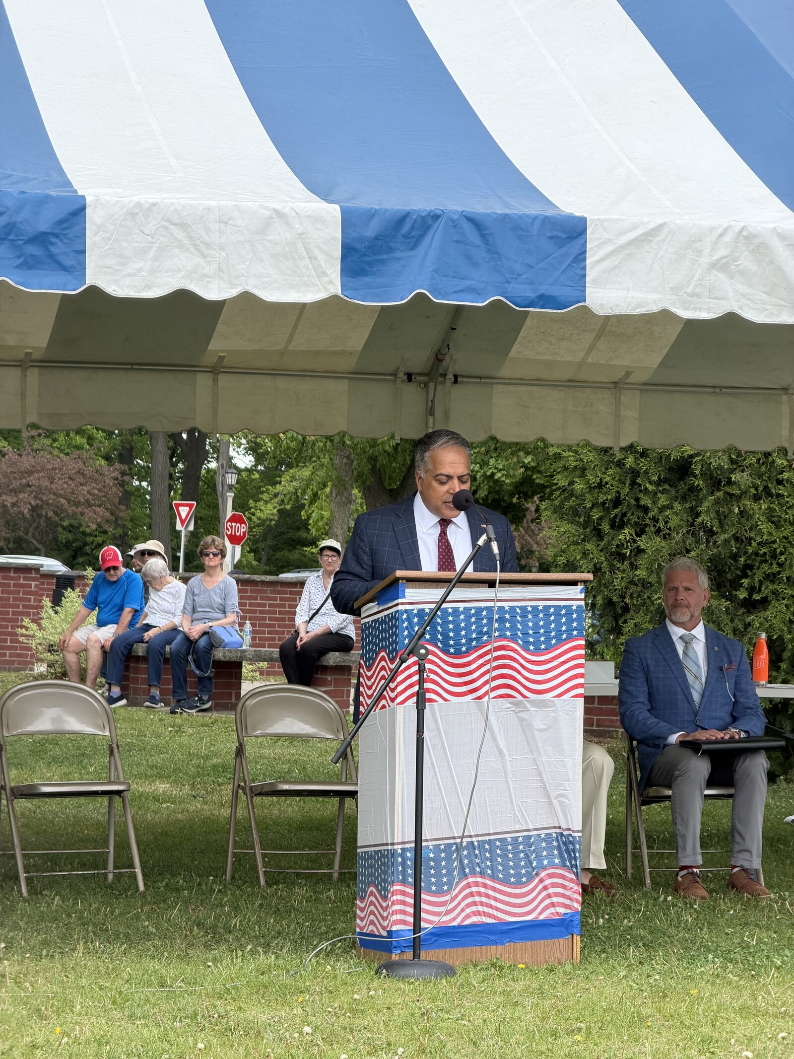 Whitefish Bay resident Anoop Prakash speaks from a podium draped in American flag bunting at the Memorial Day service, with a seated audience member visible to his right