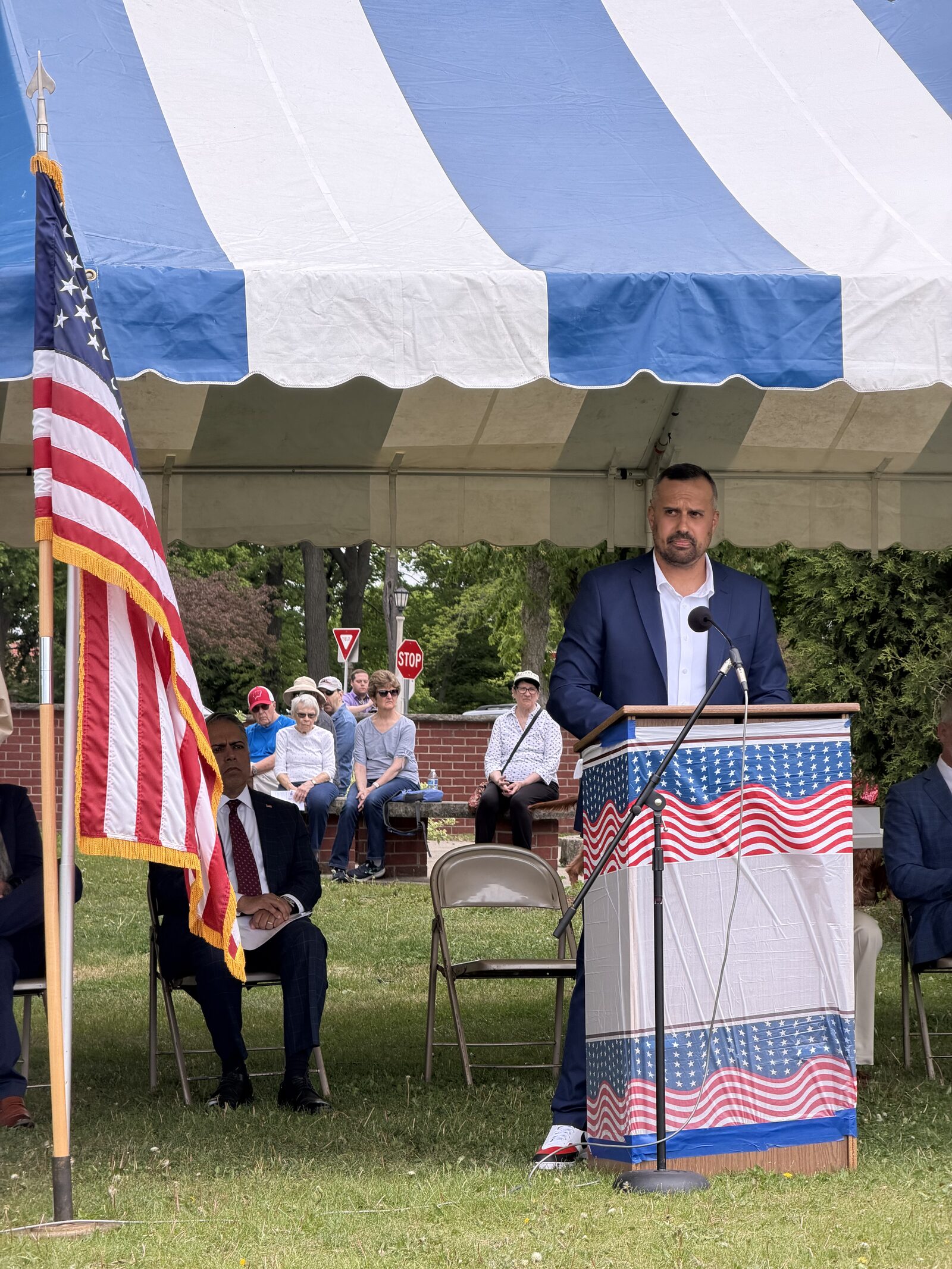 A speaker in a navy jacket addresses the Memorial Day audience from the podium, with an American flag and seated dignitaries beside him under the event tent