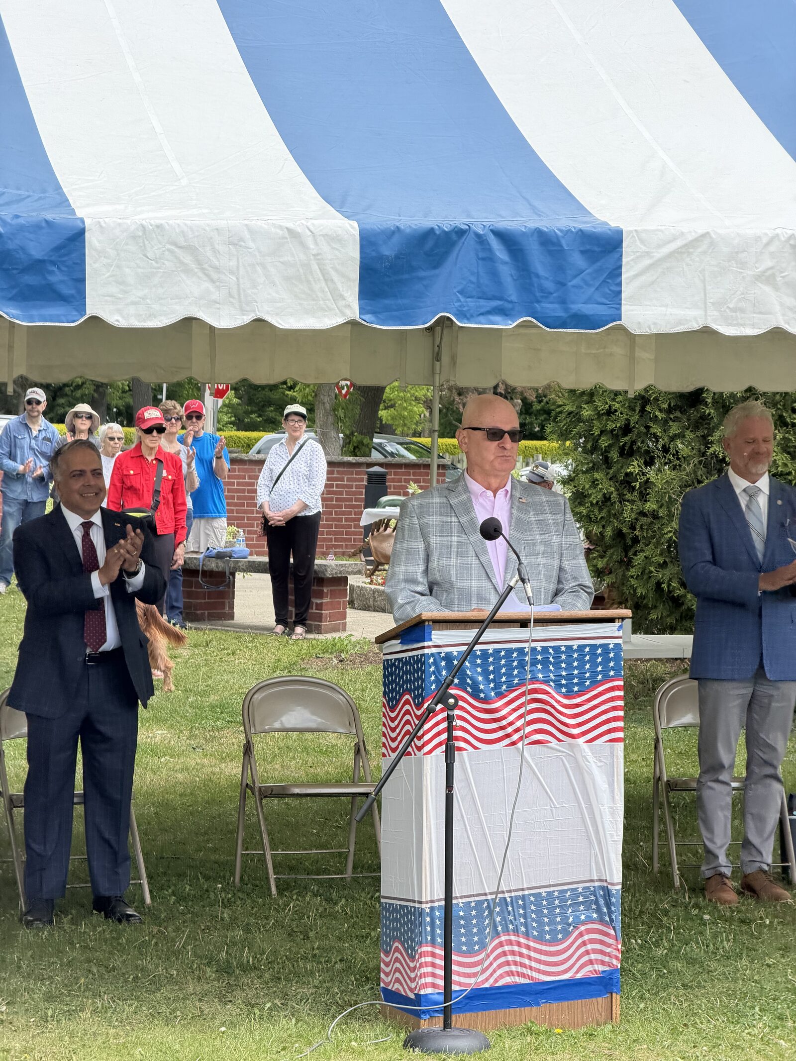 Marine Corps veteran Zach Zdroik delivers Memorial Day keynote remarks from a podium draped in American flag bunting under the blue-and-white event tent